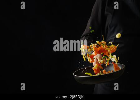 Ein professioneller Koch in schwarzer Uniform bereitet Meeresfrüchte mit Gemüse in einer Pfanne auf schwarzem Hintergrund zu. Gefrorenes Essen während des Fluges. Hotel, Restaurant Stockfoto