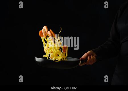 Italienische Pasta mit Garnelen in einer Pfanne auf schwarzem Hintergrund. Der Prozess des Kochens von Meeresfrüchten durch die Hände eines professionellen Küchenchefs. Gefrorenes Essen während des Fluges. Stockfoto