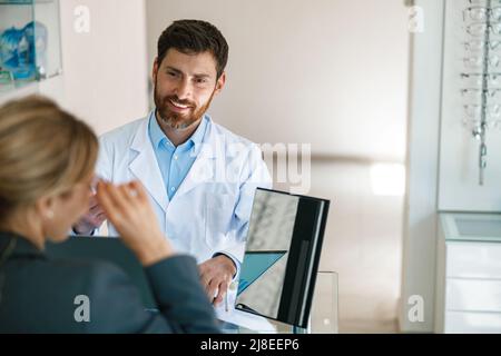 Männlicher Optiker hilft Frau, Brille im optischen Laden zu wählen, der zu ihrem Spiegel zeigt Stockfoto