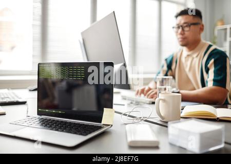 Hintergrundbild eines Laptops mit grünen Codezeilen auf dem Bildschirm im Büro von IT-Entwicklern mit Person, die am Schreibtisch arbeitet, Kopierbereich Stockfoto