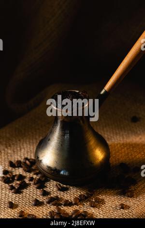 Ein alter Kupferzacke steht auf einem dunklen Hintergrund. Die Kaffeebohnen sind auf der Sackleine verstreut. Stockfoto