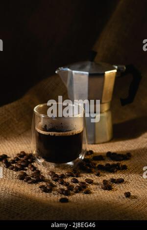 Eine durchsichtige Glastasse mit Kaffee und eine Geysir-Kaffeemaschine auf dunklem Sackleinen-Hintergrund mit Kaffeebohnen. Stockfoto