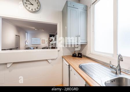 Küche mit hellem Edelstahlwaschbecken unter dem Fenster und weiß gerahmter Servierluke Stockfoto