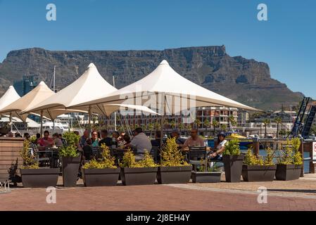 Kapstadt, Südafrika. 2022. Tafelberg und Touristen essen und trinken unter Sonnenschirmen am Wasser. Stockfoto
