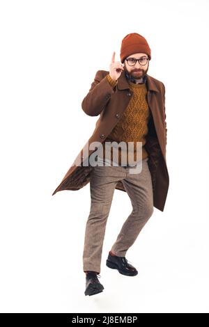 Die Studioaufnahme auf weißem Hintergrund zeigt einen eleganten Hipster mit einem Bart, der einen braunen Wintermantel trägt, mit einer Hand in der Tasche und einem Finger nach oben. Hochwertige Fotos Stockfoto
