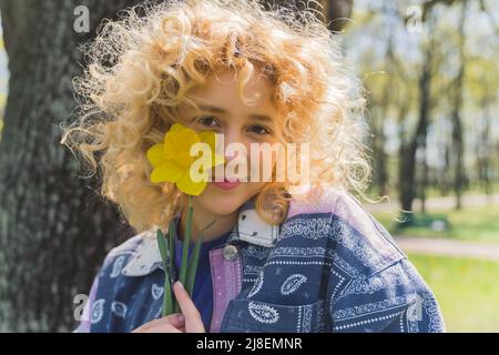 Kaukasische blonde lockige Mädchen in einem Park mit einer gelben Blume in der Nähe ihres Gesichts. Außenaufnahme. Hochwertige Fotos Stockfoto