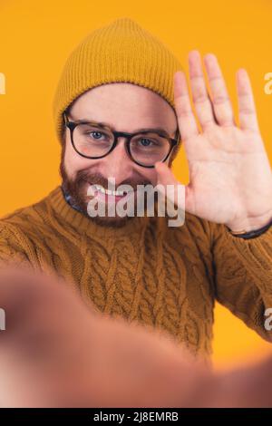 Das vertikale Studio wurde auf gelbem Hintergrund von einem bärtigen kaukasischen Mann mit einem gelben Hut und einem senfgemusterten Pullover aufgenommen, der eine Brille trug, die eine Kamera anwinkte. Hochwertige Fotos Stockfoto