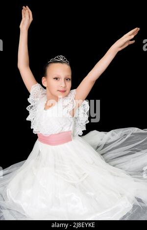 Mädchen Ballerina in weißen langen Kleid sitzt auf schwarzem Hintergrund und posiert elegant mit den Armen angehoben. Kaukasische Balletttänzerin im Alter von neun Jahren Stockfoto