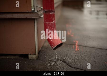 Downspout im Gebäude. Wasser fließt vom Dach auf Asphalt. Rotes Rohr zum Ablassen von Sedimenten. Stockfoto