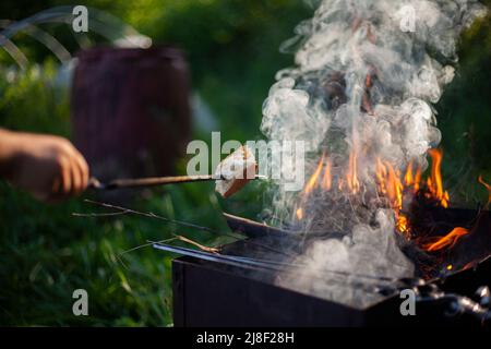 Ein Stück Brot wird auf Feuer gebacken. Picknick im Sommer im Detail. Offenes Feuer von trockenen Ästen. Kochen auf Flamme. Ruhe im Sommer. Brot auf Stock. Stockfoto