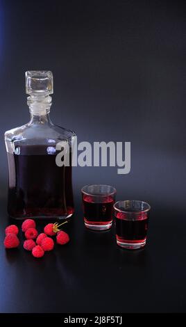 Himbeerlikör, hausgemachter Alkohol und eine Handvoll reifer Gartenbeeren auf schwarzem Hintergrund. Nahaufnahme. Stockfoto