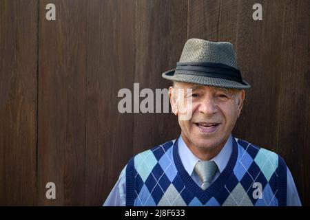 Weißer, älterer sanftmütiger Mann, der in der Nähe eines vertikalen dunkelbraunen Plankenzauns mit einem grauen Fedora-Hut lächelt Stockfoto