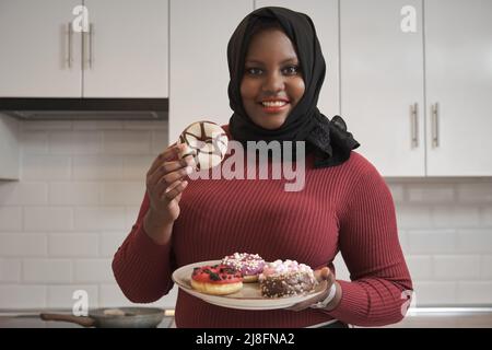 Porträt einer jungen muslimischen Frau, die einen Donuts-Teller in der Hand hält und lächelt. Stockfoto