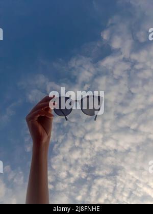 Frau Hand mit Sonnenbrille auf dem Hintergrund des blauen Himmels. Blick auf den Himmel durch Sonnenbrille Stockfoto