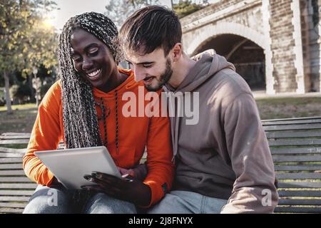 Multiethnisches Paar junger Menschen, die ein digitales Tablet verwenden, um Inhalte zu durchsuchen, die auf einer Bank im Park sitzen – Lifestyle-Konzept von biraci Stockfoto