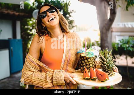 Eine niedliche junge Frau bringt ein Tablett mit Obst auf die Terrasse, Ananas- und Wassermelonendesserts zum Tisch der Freunde, während sie die Kamera anlächelt. Stockfoto