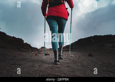 Rückansicht einer Wanderfrau, die auf dem Lavastein des Vulkans Ätna in Sizilien mit Trekkingstöcken, roter Weste und Rucksack spazierengeht - Wanderlust Lifestyle c Stockfoto