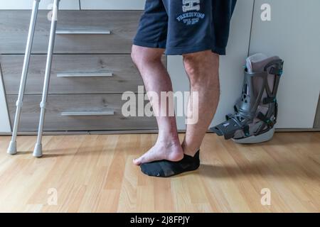 Europäischer Mann nach Achillessehnenriss Operation ist wieder zu Hause mit Moon Boot speziellen physiotherapeutischen Schuh und Krücken für die Erholung zu Hause gegen Stockfoto