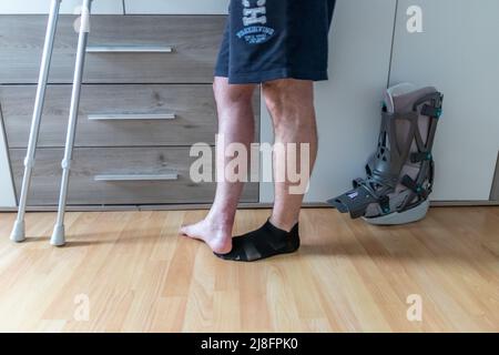 Europäischer Mann nach Achillessehnenriss Operation ist wieder zu Hause mit Moon Boot speziellen physiotherapeutischen Schuh und Krücken für die Erholung zu Hause gegen Stockfoto