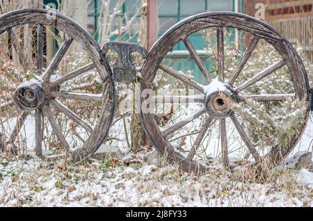 Ein paar Räder eines alten Wagens, die sich an einen Zaun lehnen, der in der Vergangenheit schon seit Meilen benutzt wurde. Holzräder des Wagens mit Schnee bedeckt. Stockfoto