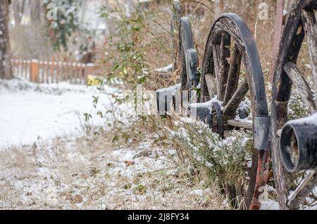 Ein paar Räder eines alten Wagens, die sich an einen Zaun lehnen, der in der Vergangenheit schon seit Meilen benutzt wurde. Holzräder des Wagens mit Schnee bedeckt. Stockfoto