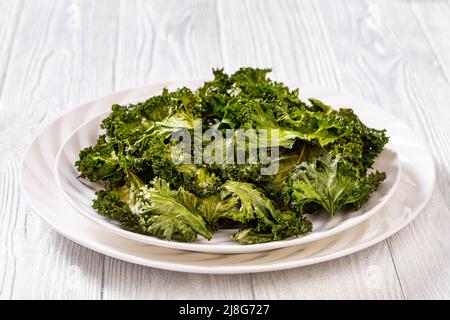 kale-Chips auf weißem Teller auf weißem strukturiertem Tisch, horizontale Ansicht von oben Stockfoto