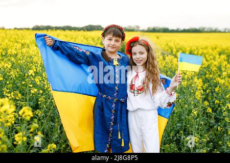 Betet für die Ukraine. Zwei niedliche Mädchen halten in Händen eine blau-gelbe ukrainische Nationalflagge in der Mitte eines Rapsfeldes Stockfoto