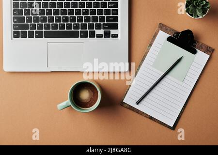 Draufsicht auf Laptop-Tastatur, Tasse Kaffee, grüne Hauspflanze in kleiner Blumentopf, Klemmbrett mit Notizbuch und Stift auf beigem Schreibtisch Stockfoto