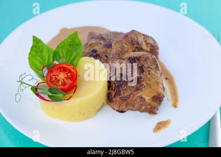 Fleischbällchen mit Kartoffelpüree auf einem weißen Teller. Fleisch in saurer Rahmsoße. Tomaten und Basilikum. Essen im Restaurant. Mittagspause. Medaillons. Stockfoto