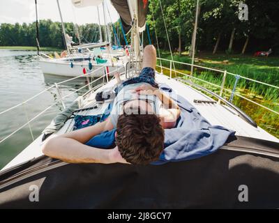 Erwachsener Mann, der an einem sonnigen Sommertag auf einem Segelbootdeck liegt und sich während seines Urlaubs sonnen hat. Yacht liegt an einem hölzernen Pier an einem Seeufer Stockfoto