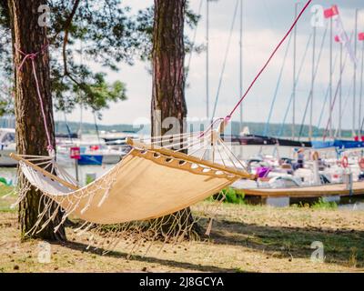 boho-Stil weiße Leinenhängematte, die zwischen Pinien an einem Seeufer mit Segelbooten Hafen im Hintergrund hängt Stockfoto