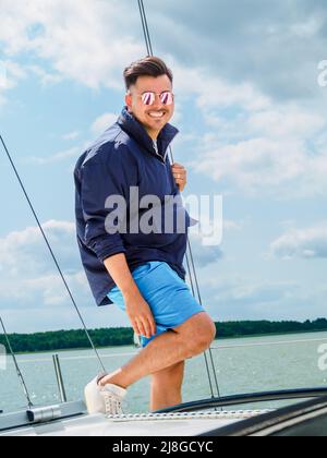 Der junge, schwarze, lachende Mann trägt eine Sonnenbrille und ein nautisches Tuch und steht auf einem Segelboot während seiner Segelreise im Sommer Stockfoto