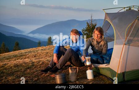 Familie paar Touristen kochen leckeren Kaffee auf Herd Brenner der Ballonpatrone mit Gas für Wanderungen im Zeltlager auf dem Bergwiese. Neblige Berge und grauer Himmel hinter ihnen in die Ferne. Stockfoto