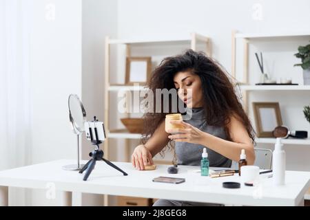 Fröhlich gebräunt schöner lockiger lateinischer Beauty-Blogger in Casual Top erzählt von Körpercreme im Heim-Interieur. Space Mockup Banner kopieren. Influencer-Rekord Stockfoto