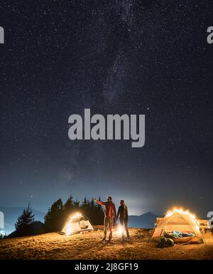 Mann, der in der Nähe von beleuchteten Zelten steht, die Hand seiner Freundin hält und sie auf die Landschaft vor ihm zeigt. Nachtcamping auf Bergwiese unter Sternenhimmel mit Milchstraße. Konzept des Campings. Stockfoto