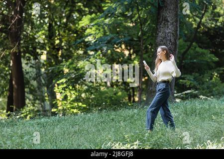 Glückliche junge Frau, die während eines Videoanrufs auf dem Smartphone die Hand schwenkt, während sie im Park läuft Stockfoto