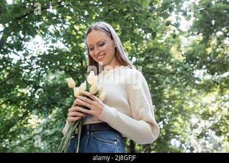 Low-Angle-Ansicht einer glücklichen Frau in einer Brille, die Tulpen im grünen Park hält Stockfoto