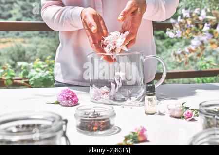 Frau bereitet Rosenwasser mit rosa Rosenblüten in einer Glasschüssel zu. Hautpflege und Spa, natürliche Schönheitsbehandlungen, hausgemachte Kosmetik. Stockfoto