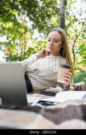 Nachdenkliche Frau in einer Brille, die einen Pappbecher hält und auf dem Laptop auf der Decke im Park schaut Stockfoto
