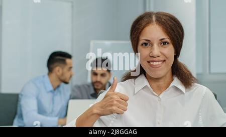 Lächelnde, freundliche Geschäftsfrau, die die Kamera anschaut und wie Daumen nach oben zeigt. Enthusiastisch glücklich erfolgreiche Führerin Managerin sitzt in modern Stockfoto