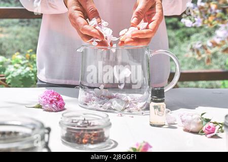 Frau bereitet Rosenwasser mit rosa Rosenblüten in einer Glasschüssel zu. Hautpflege und Spa, natürliche Schönheitsbehandlungen, hausgemachte Kosmetik. Stockfoto