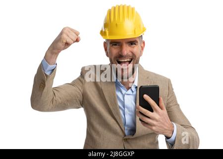 Schöner bärtiger Ingenieur in Anzug mit Helm, der Telefon hält, mit Arm in der Luft lacht und vor weißem Hintergrund jubelt Stockfoto