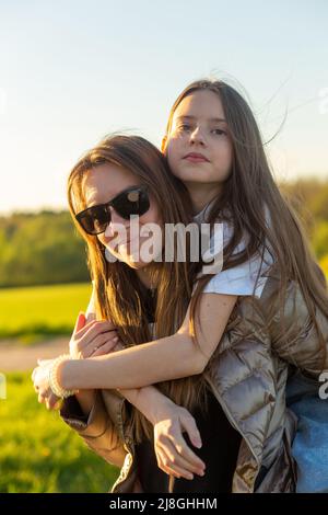 Verspielte Mutter gibt Tochter Piggy zurück Fahrt auf dem grünen Feld. Beide lachen und sehen glücklich aus. Frühling im Wald Hintergrund. Nahaufnahme. Stockfoto