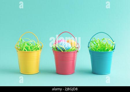 Osterfesttagskarte mit bunten Eiern in gelben, blauen und roten dekorativen Eimern auf pastelltürkisfarbenem Hintergrund. Konzept für die Eiersuche. Stockfoto