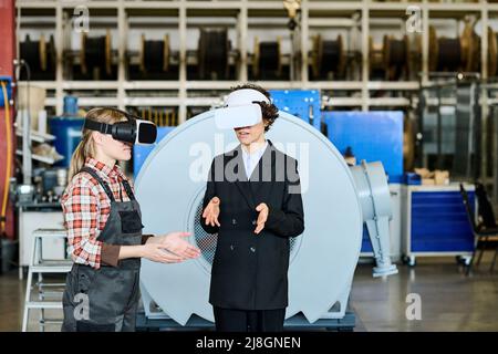 Zwei junge Ingenieurinnen in vr-Headsets diskutieren 3D Modelle ihres Produkts, während eine von ihnen eine Präsentation bei einem Meeting vorstellt Stockfoto