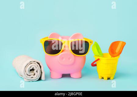 Sommerferienkonzept. Rosa Sparschwein-Geldbank mit Strandtuch, Sonnenbrille und Strandeimer auf blauem Hintergrund Stockfoto