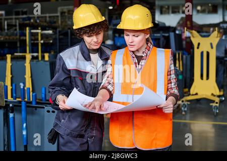 Zwei Arbeiter der modernen Fabrik tragen Schutzhelme und Arbeitskleidung und betrachten die Skizze auf blauem Druck, während einer von ihnen sie erklärt Stockfoto
