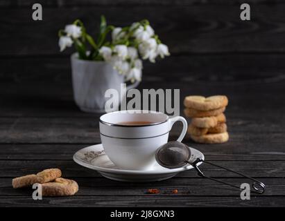 Eine Tasse Tee mit Keksen und Teesieb auf dunklem Hintergrund mit bunten weißen Schneeglöckchen Stockfoto