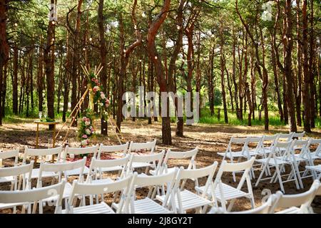 Wunderschöne böhmische Tipi-Bogendekoration auf der Hochzeitsfeier im Freien im Pinienwald mit Kegeln. Stühle, floristische Blumenkompositionen aus Rosen Stockfoto