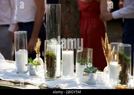Hochzeitsfeier Bankett im Freien im Wald. Esstisch im Boho-Stil mit Kerzen, weißem Tuch, Blumen, serviert mit Tellern, Geschirr dekoriert Stockfoto